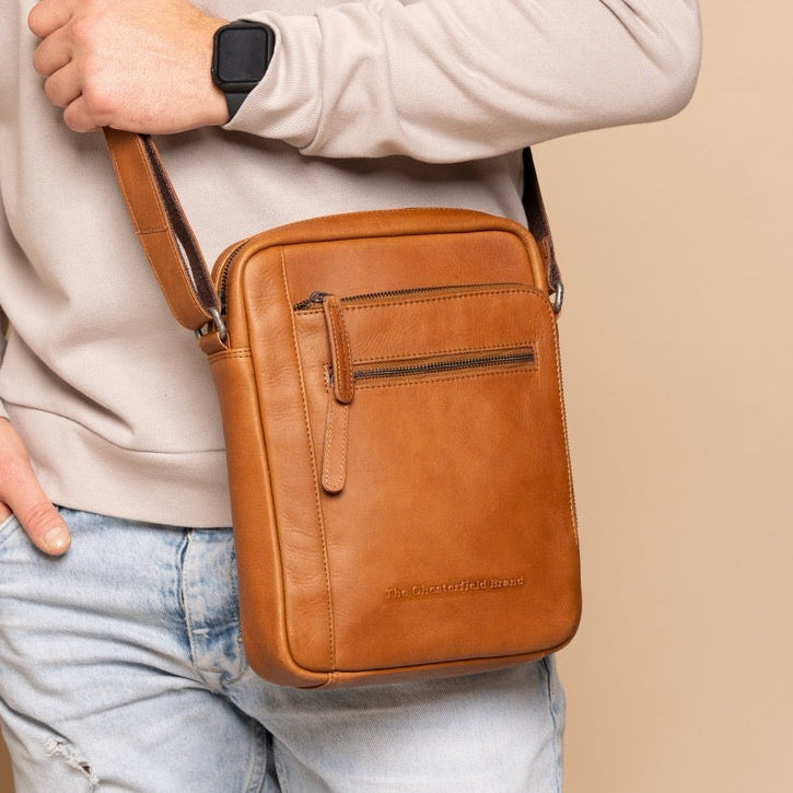 Person wearing a beige sweater and light blue jeans with a cognac leather crossbody bag against a beige wall.