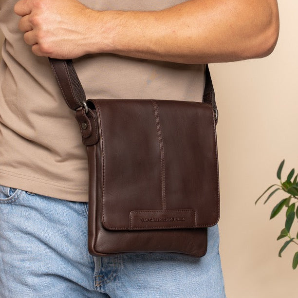 Man wearing a brown leather shoulder bag against a beige wall with a plant in the background