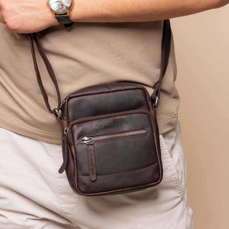 Person holding a brown leather crossbody bag against a beige background