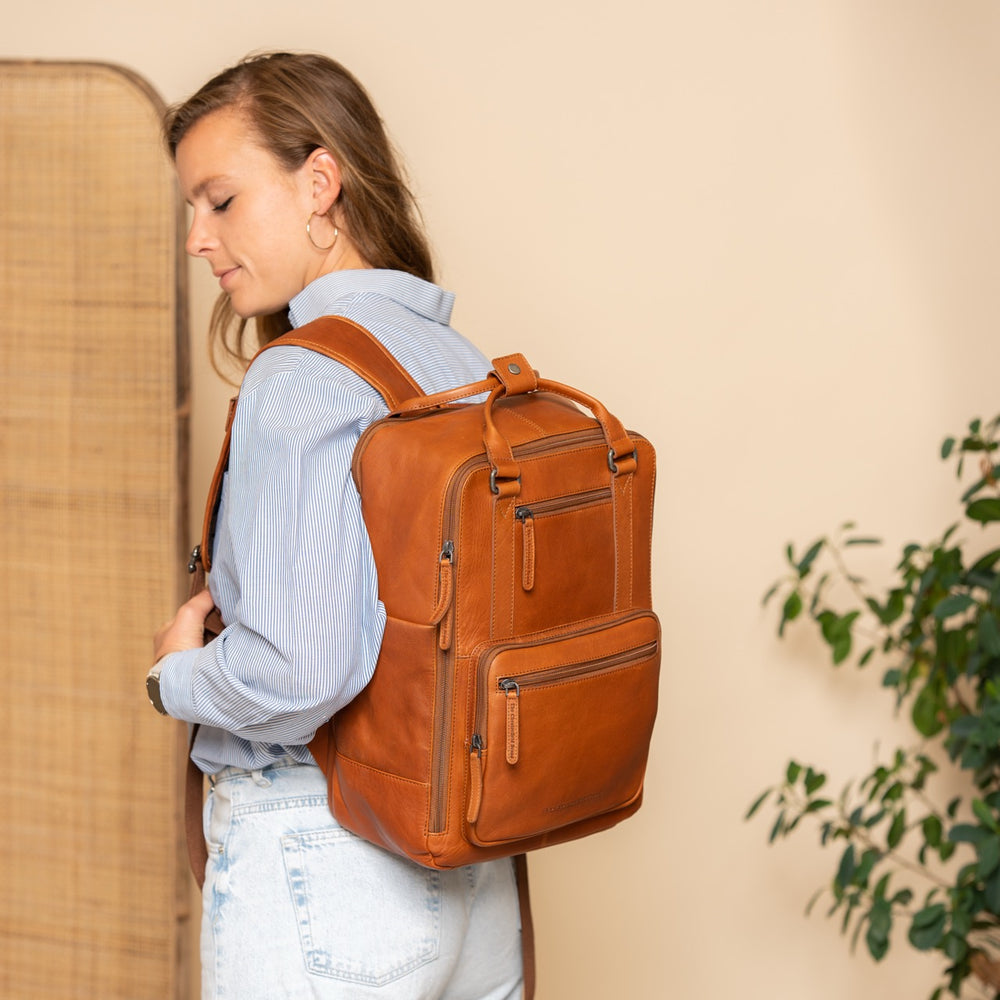 Person wearing a cognac leather backpack against a beige wall with a plant in the corner.