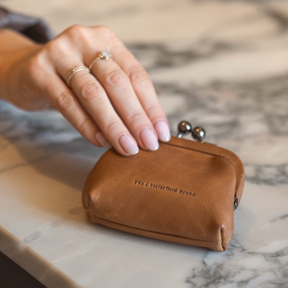 Cognac leather coin purse on a marble surface with a hand reaching towards it.