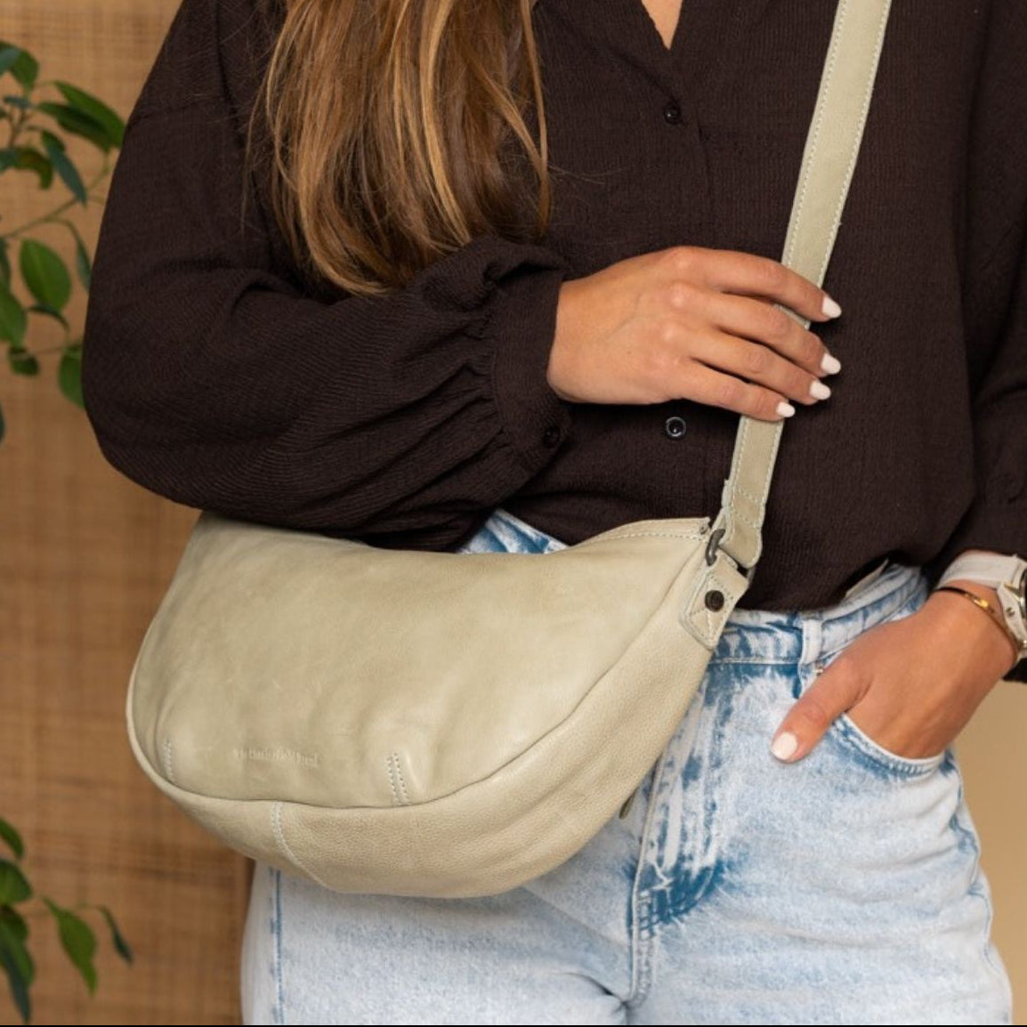 Person wearing a brown shirt and light blue jeans with a soft green crossbody bag, standing against a neutral background.