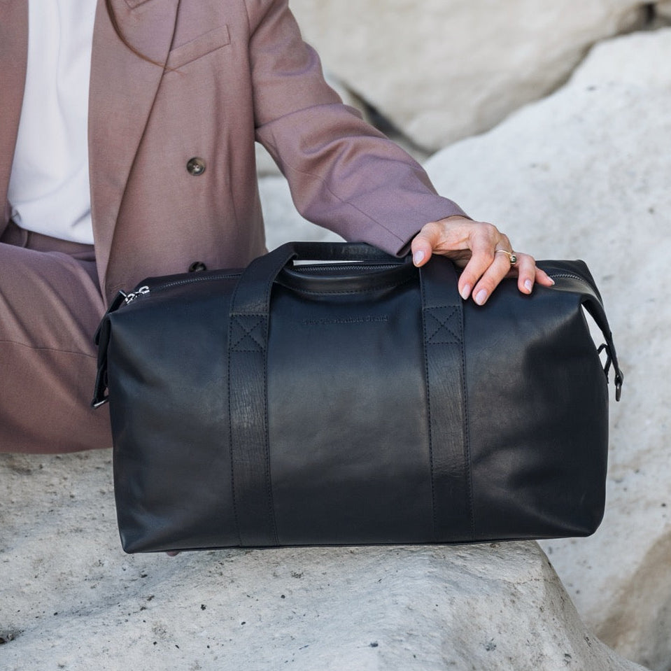 Person holding a black leather duffel bag against a rocky background.