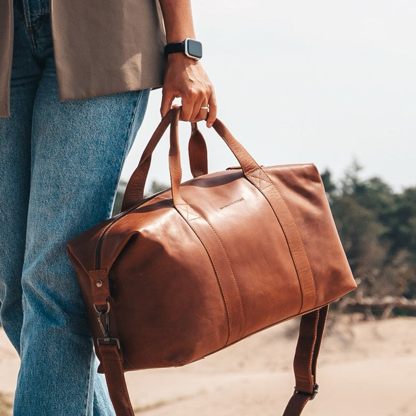 Person holding a cognac leather bag outdoors with a forest background.