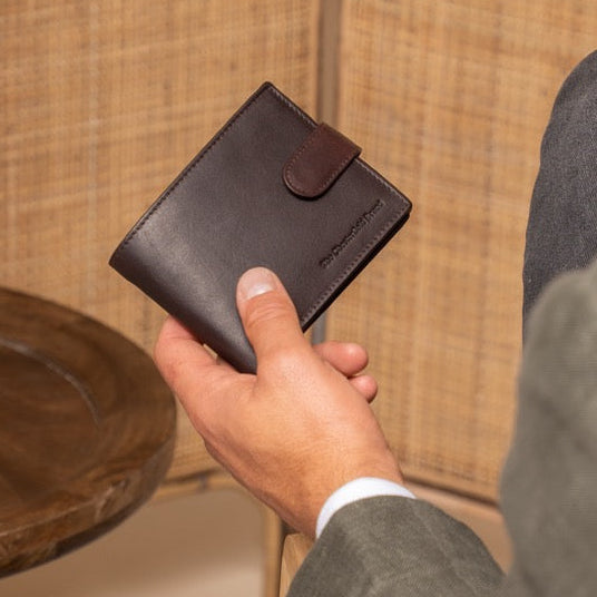 Man holding a brown leather wallet next to a vase on a wooden table against a woven wall.