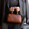 Person holding a cognac leather handbag wearing a gray coat indoors.
