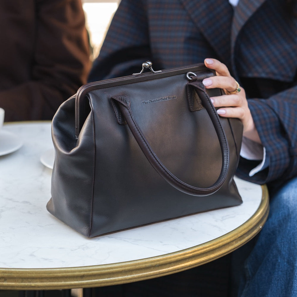 Person holding a brown leather handbag at a cafe table with a cup of coffee.