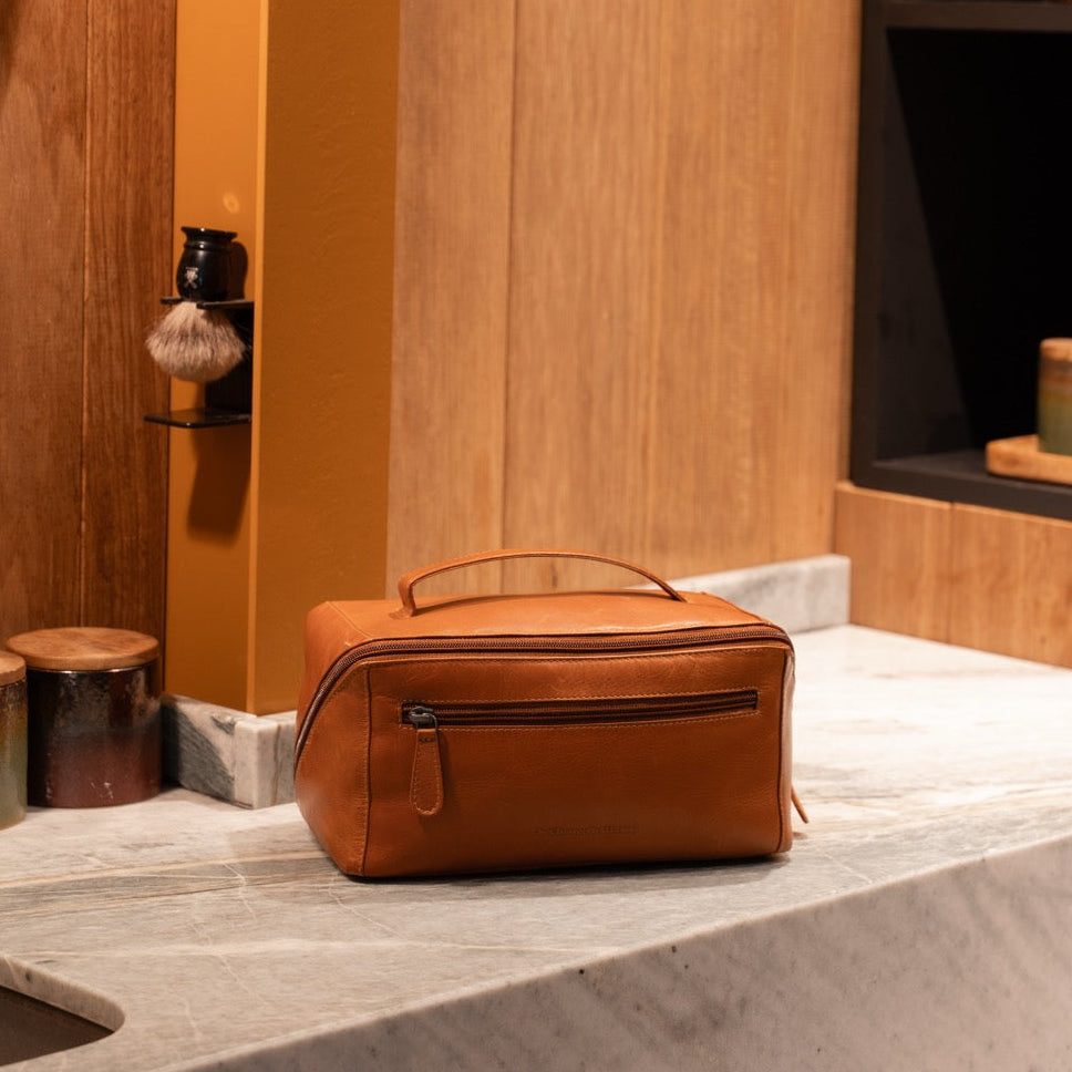Cognac leather toiletry bag on a marble surface with wooden wall and decorative items in the background