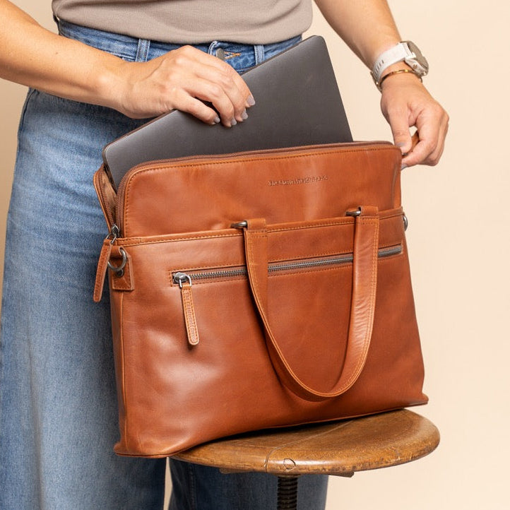 Person holding a cognac leather bag with a laptop inside, standing next to a stool.
