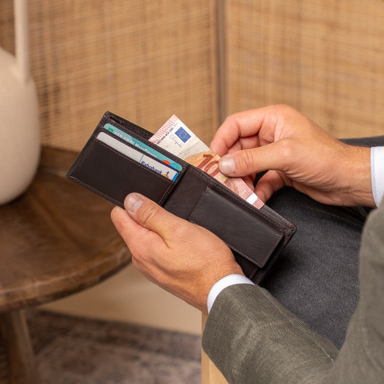 Man in a suit sitting and looking into a brown wallet with money and cards.