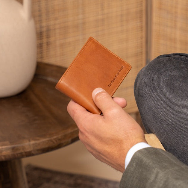 Person holding a cognac leather wallet in a room with a vase on a table.