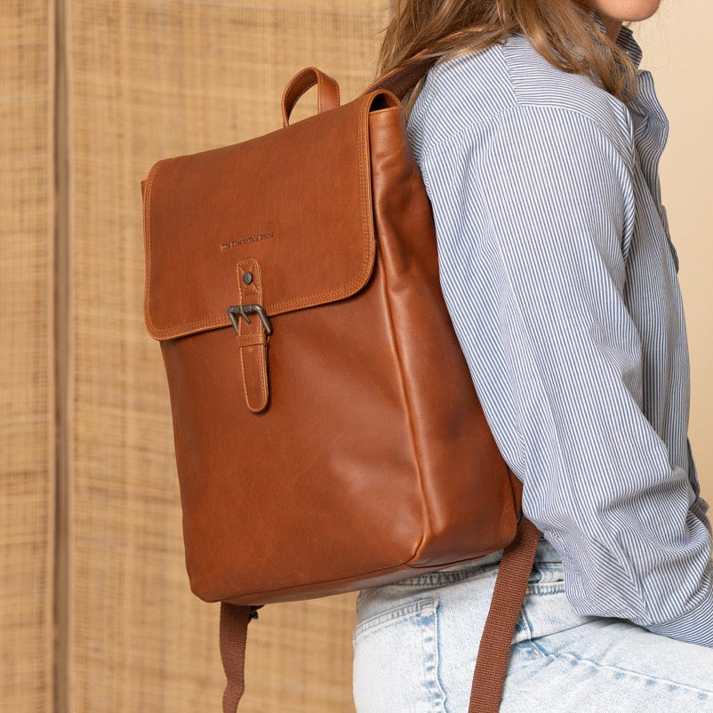 Woman wearing a cognac leather backpack sitting on a stool against a beige wall.