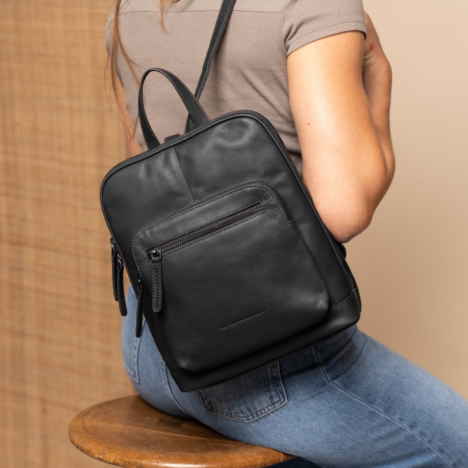 Person wearing a black backpack sitting on a wooden stool against a beige wall.