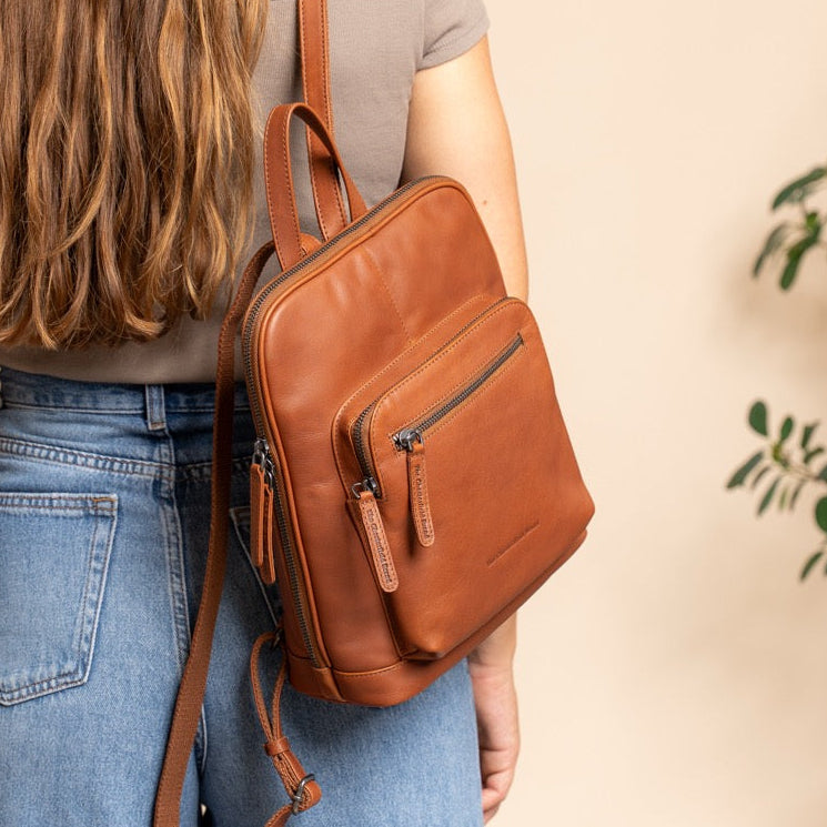 Woman wearing a cognac leather backpack against a beige wall with a plant in the corner.