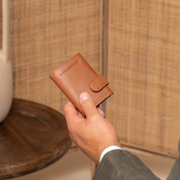 Man holding a cognac leather wallet next to a white vase on a wooden table.