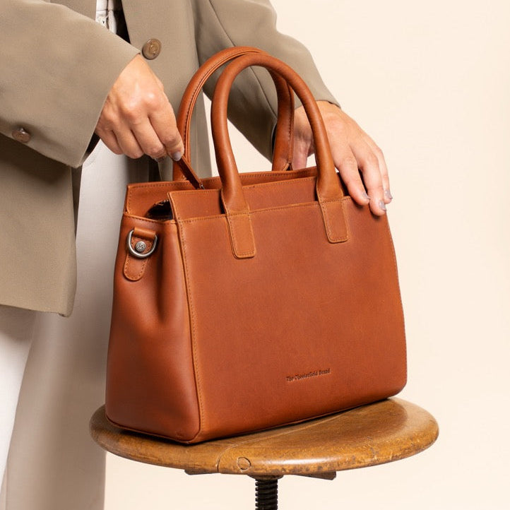 Person holding a cognac leather handbag on a wooden stool with a neutral background