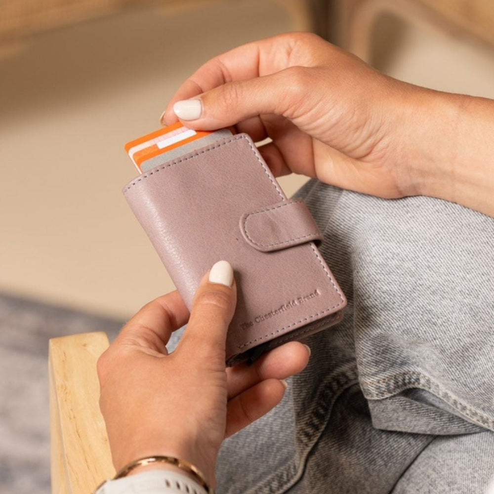 Person holding a small lavender lila wallet with an orange card inside, sitting on a wooden chair.
