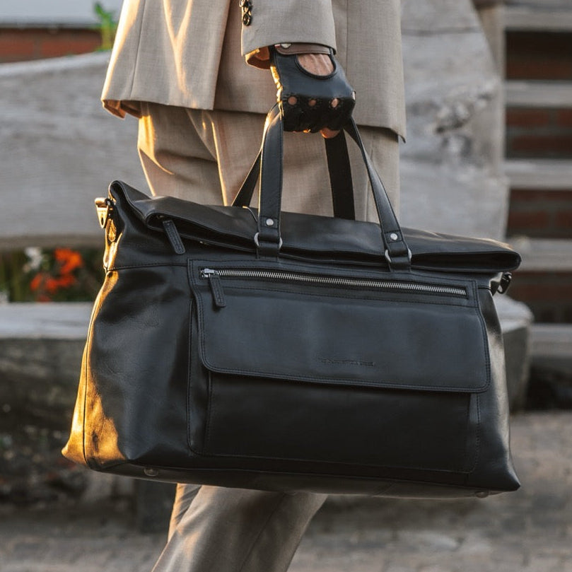 Person in beige suit holding a black leather weekender bag on a cobblestone street.