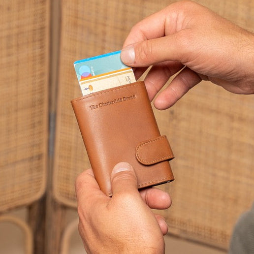 Man in a suit holding a cognac leather wallet with a card inside, sitting on a chair.