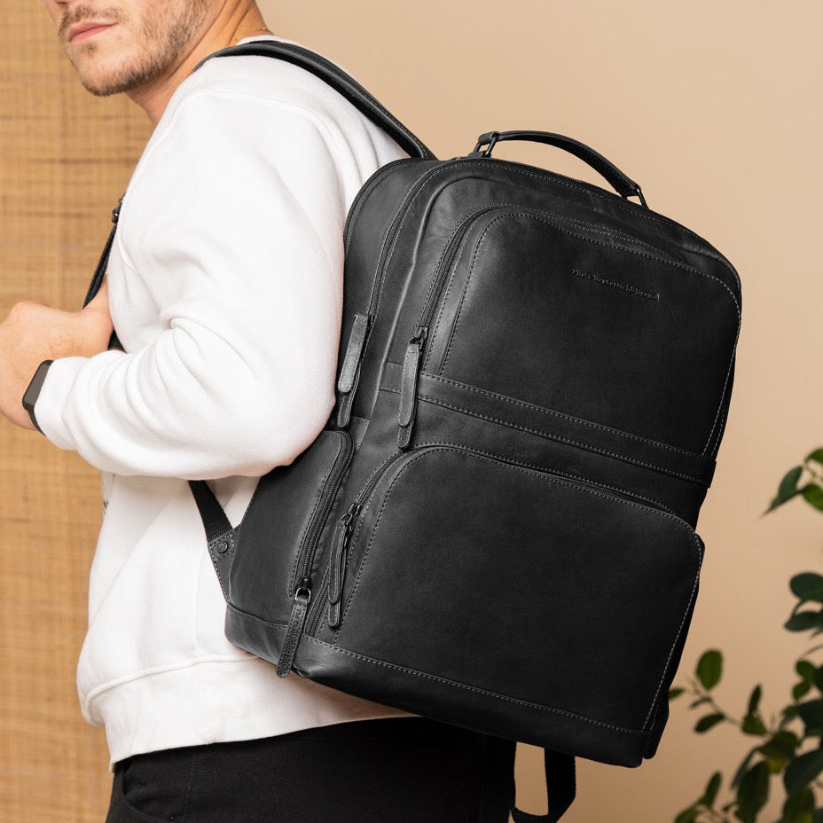 Man wearing a black backpack against a beige wall with a plant in the corner