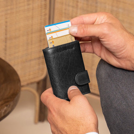 Man sitting on a chair holding a black wallet with a card, with a vase and woven wall in the background.