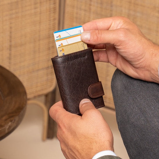 Man sitting on a chair holding a wallet with a card, with a vase and woven wall in the background.