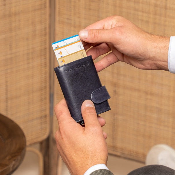 Man in a suit holding a navy leather wallet with a card in it, sitting in a room with a wooden table and vase.