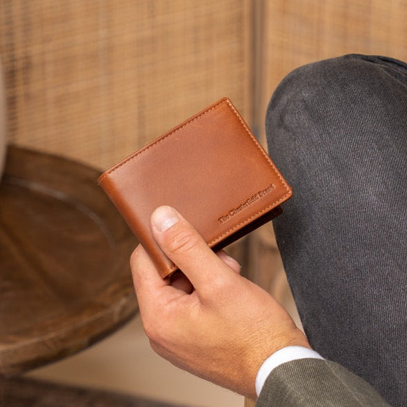 Man holding a cognac leather wallet in a room with a vase and chair.
