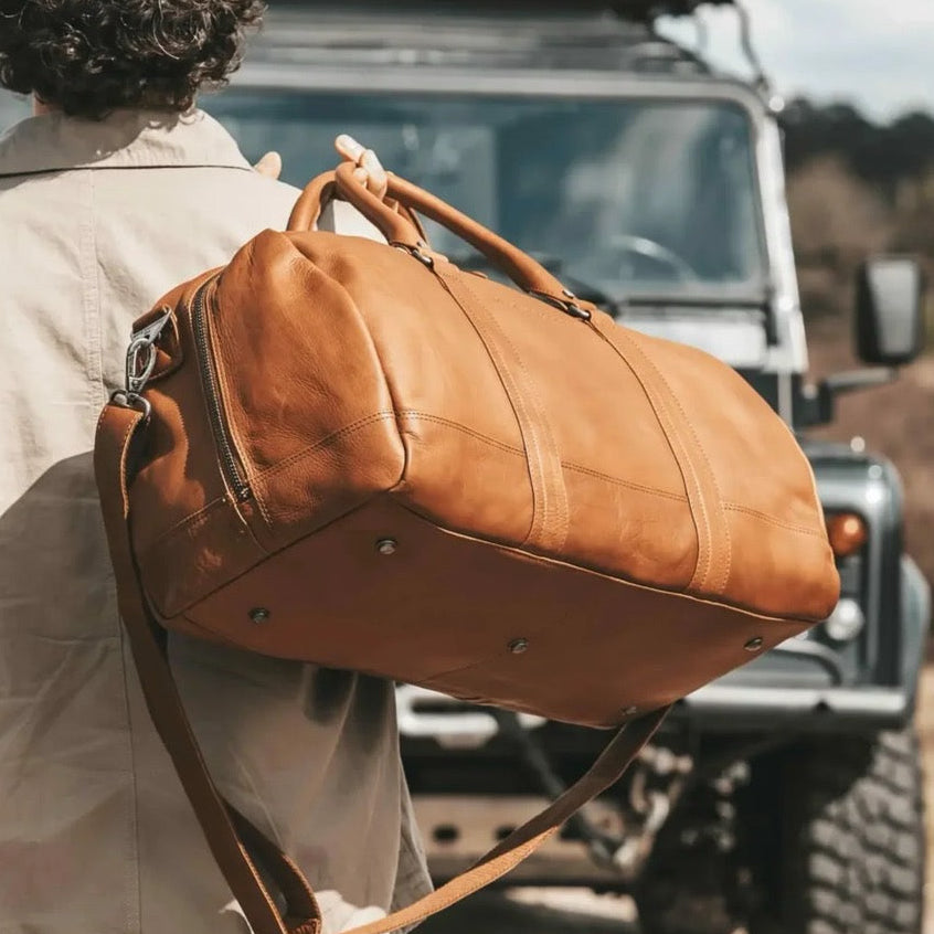 Person with a cognac leather bag walking towards an off-road vehicle in a natural setting.