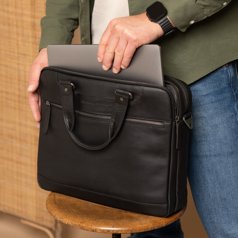 Person holding a black leather laptop bag on a wooden stool with a neutral background