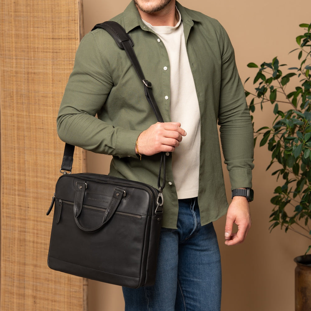 Man holding a black leather laptop bag against a beige wall with a plant in the corner