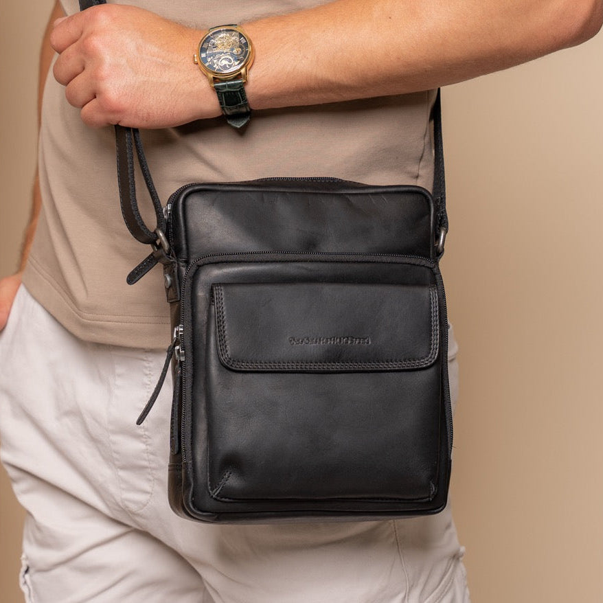 Person holding a black leather crossbody bag with a beige background