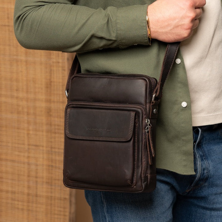 Man holding a brown leather bag against a beige wall with a plant in the background