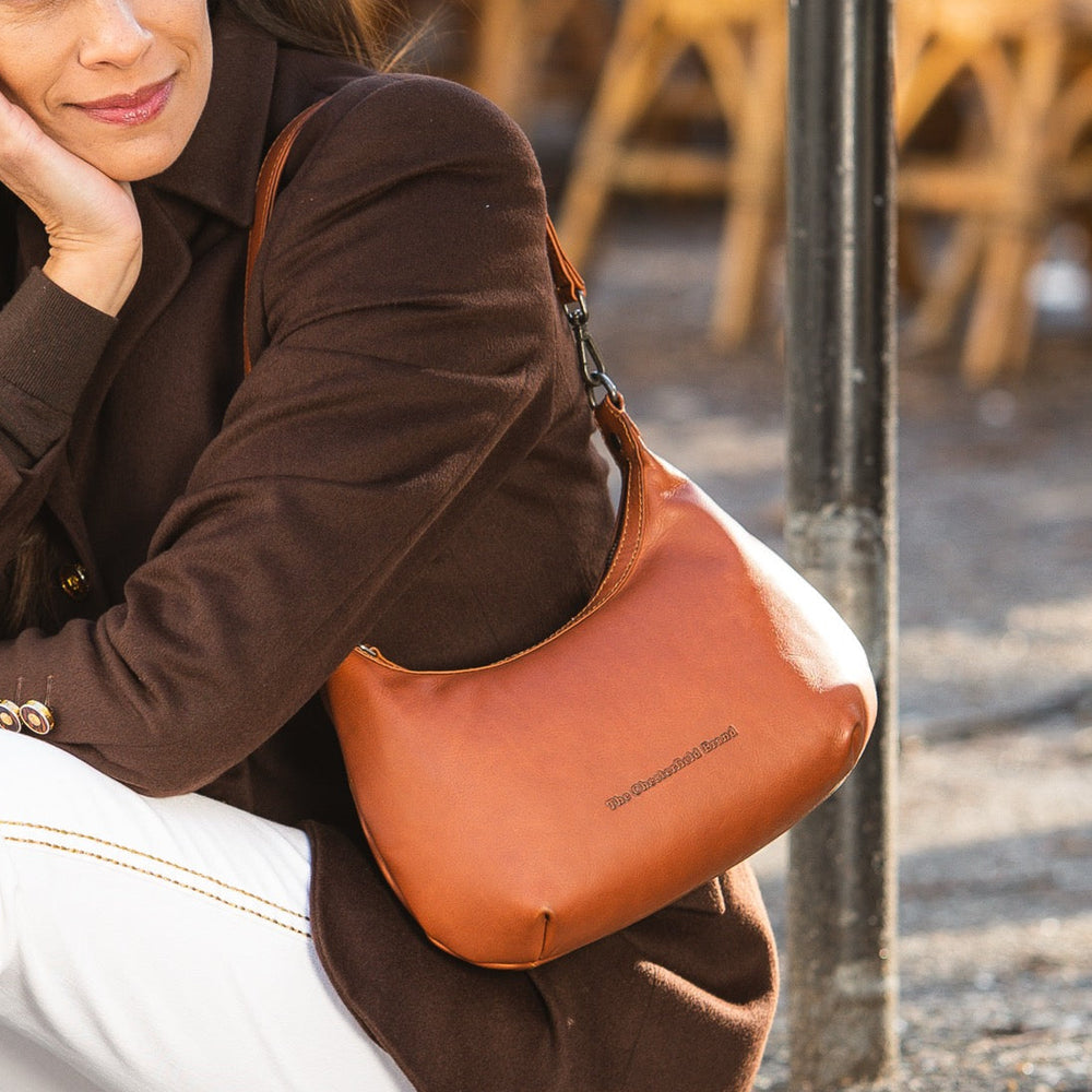 Woman sitting outdoors wearing a brown coat and holding a cognac leather handbag.