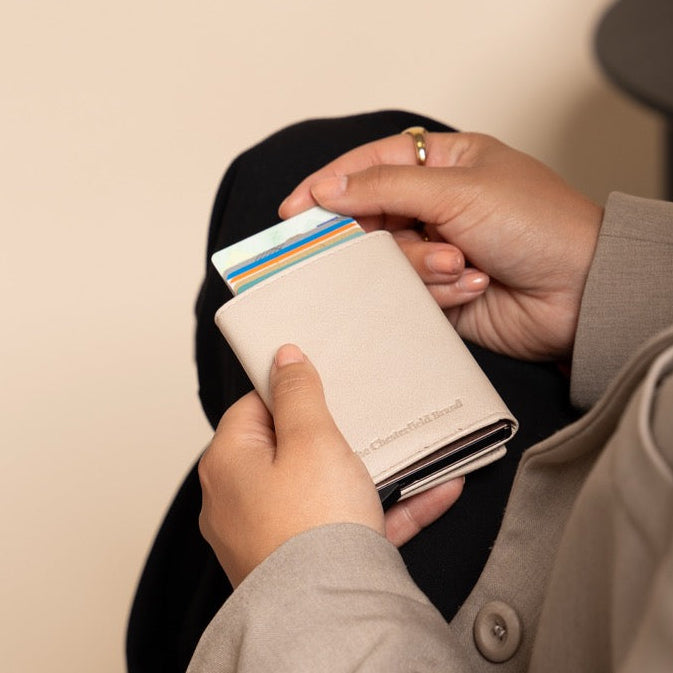 Person sitting on a chair holding a beige wallet with cards, wearing a beige coat.