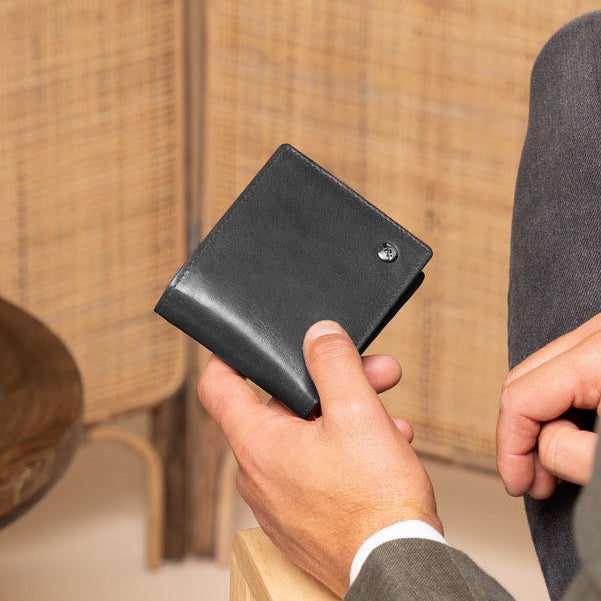 Man holding a black leather wallet in a room with wooden furniture and a woven wall.
