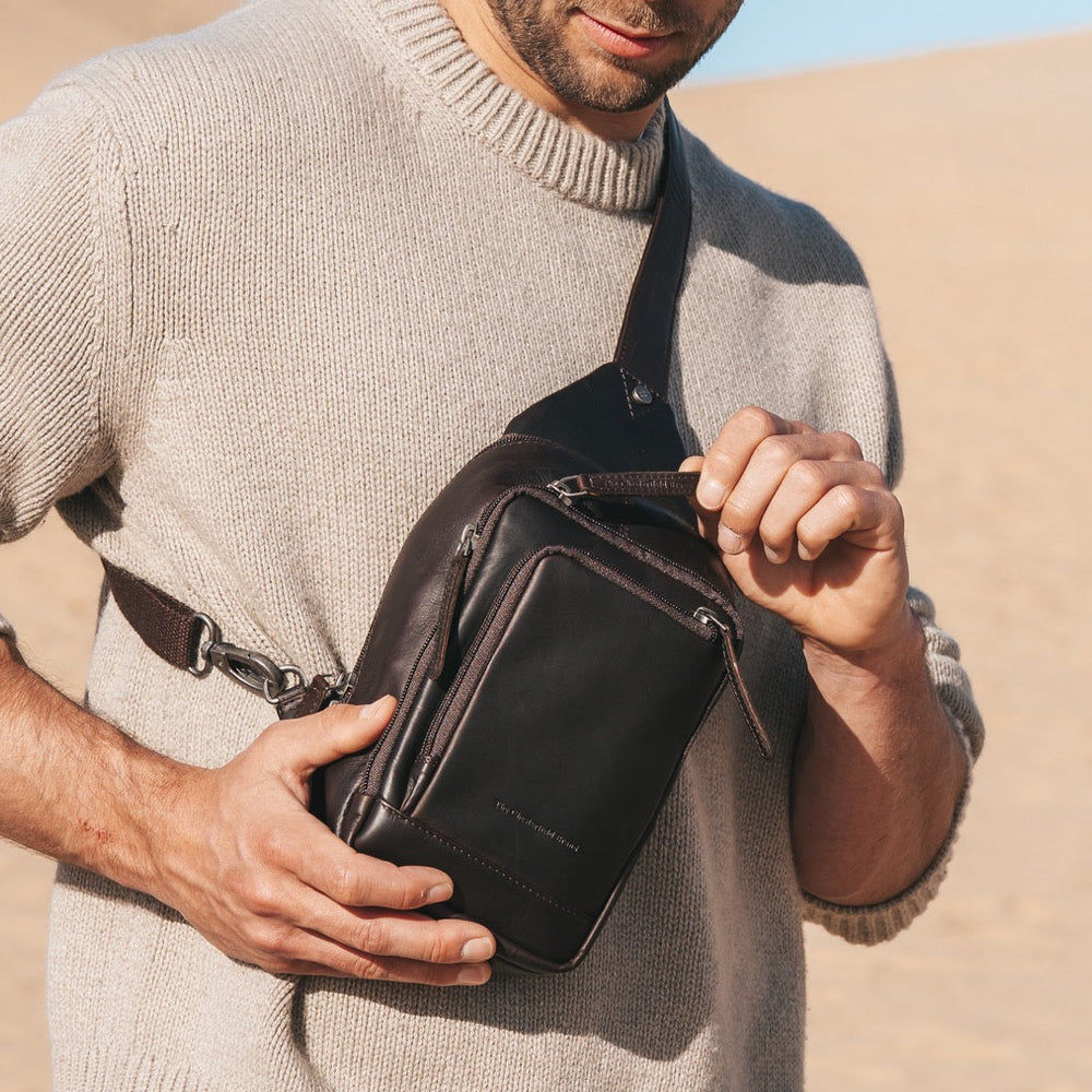 Man in beige sweater holding a brown leather sling bag in a desert setting