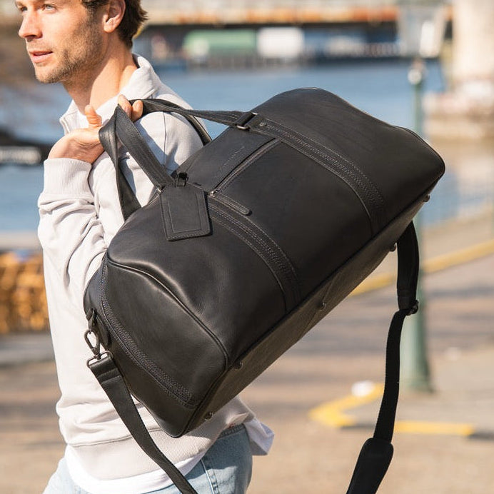Man carrying a black duffel bag outdoors near water and buildings