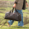 Person holding a brown leather duffel bag in a natural setting