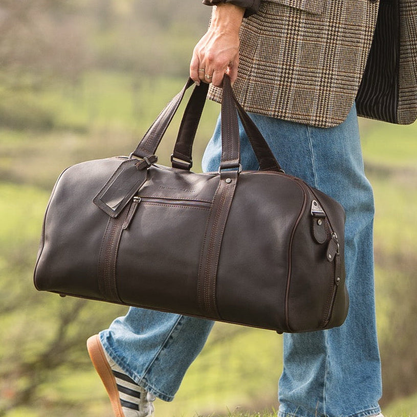 Person holding a brown leather duffel bag in a natural setting