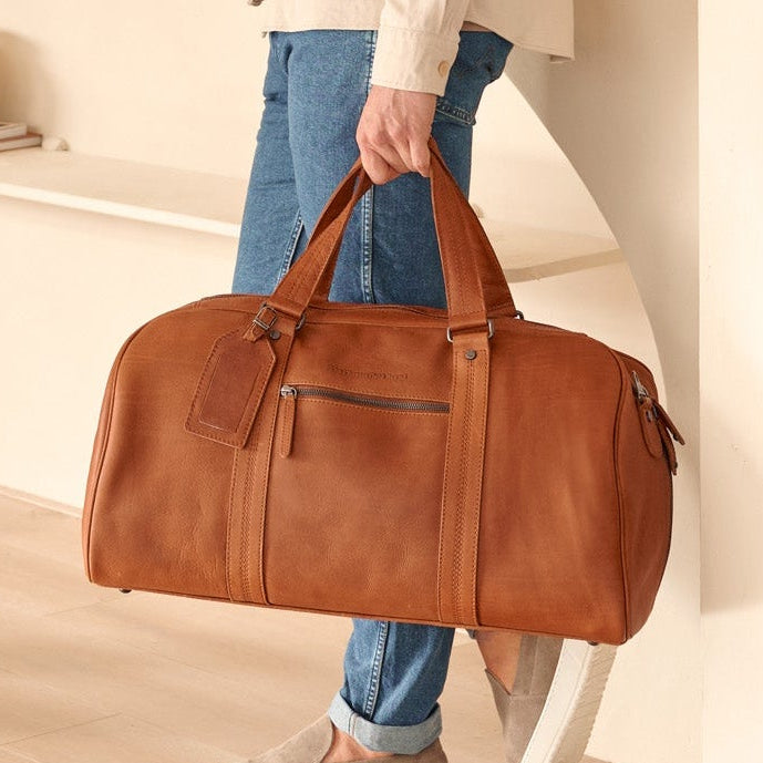 Two people standing in a modern living room with one holding a cognac leather weekender bag.