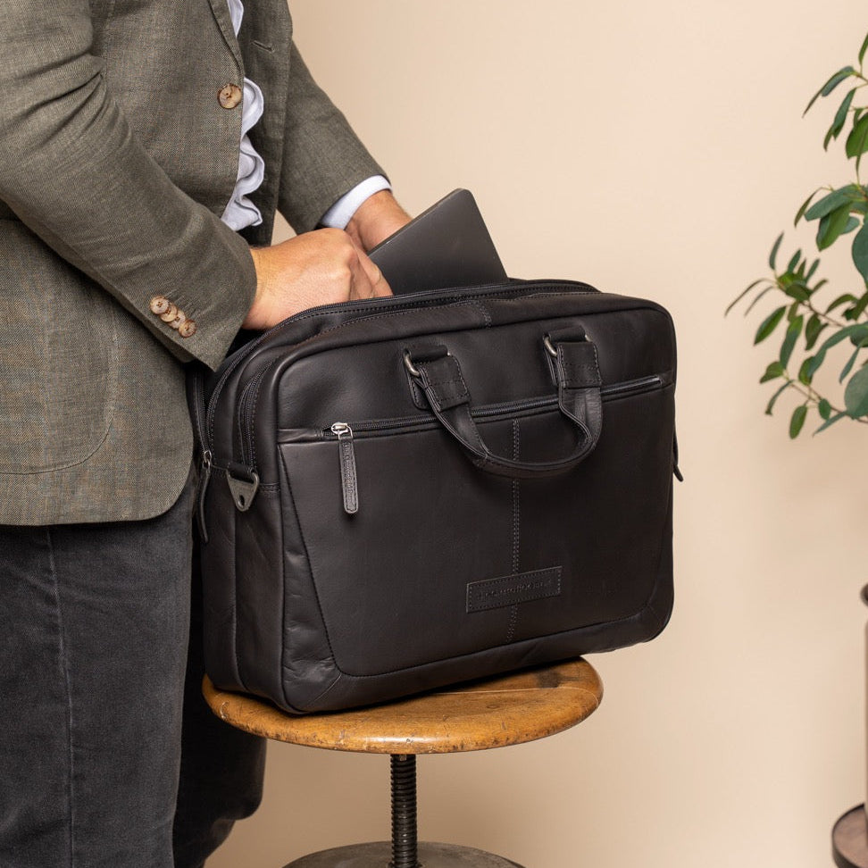 Person holding a black leather briefcase against a beige wall with a plant in the corner.