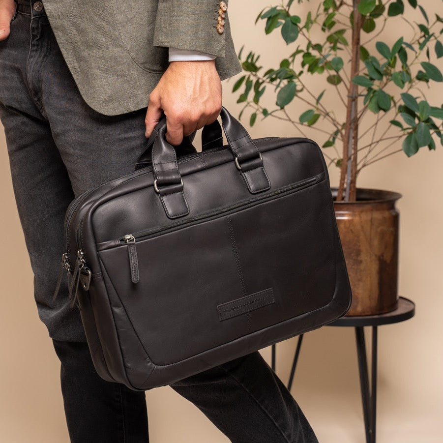 Person holding a black leather briefcase in an indoor setting with a plant in the background.