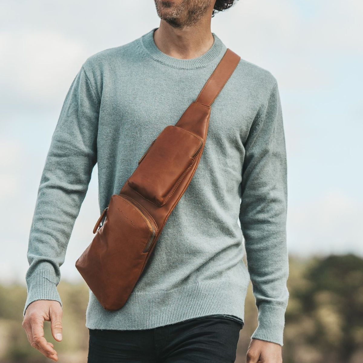 Man walking outdoors with a cognac crossbody bag, smiling