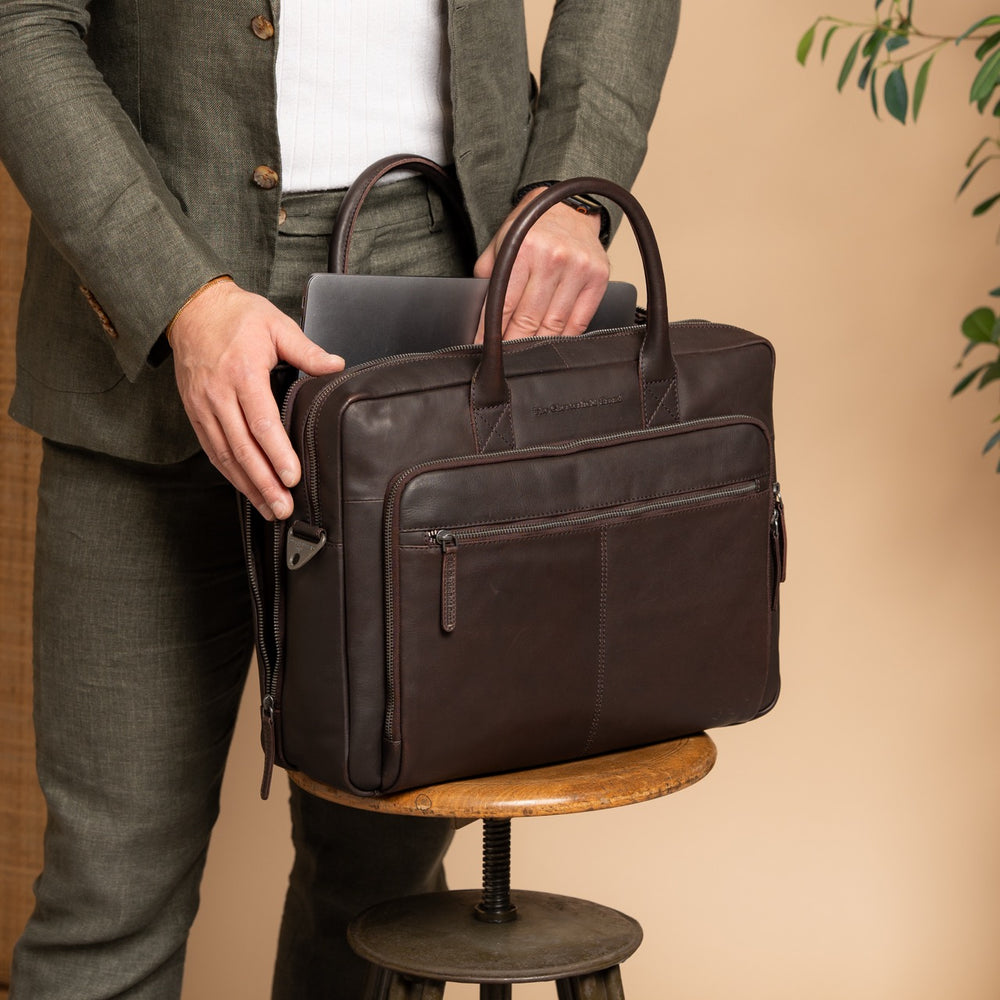 Person taking a laptop out of a brown leather briefcase on a wooden stool against a beige background