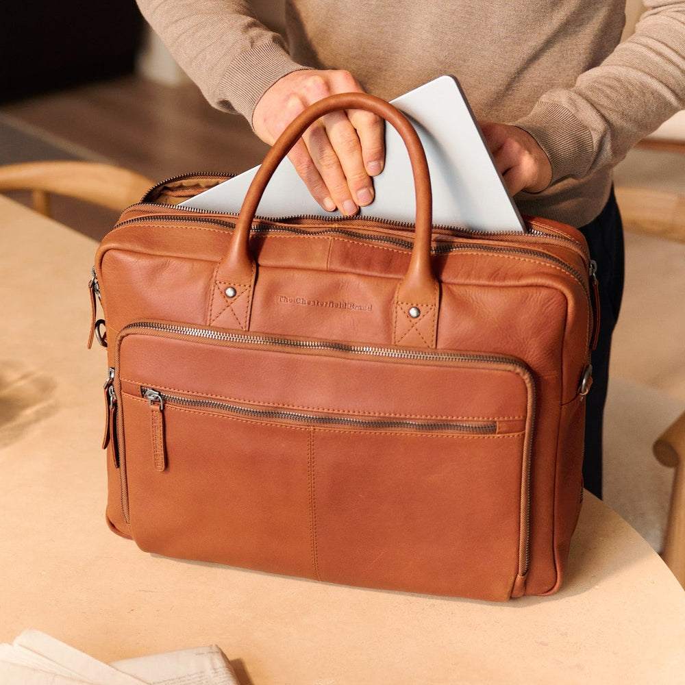Person holding a cognac leather laptop bag on a table with a laptop in the bag.