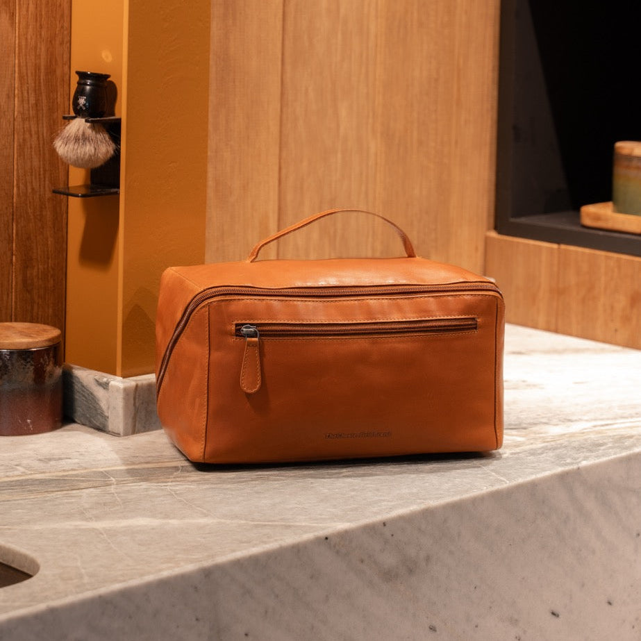 Cognac leather toiletry bag on a bathroom counter with wooden walls and decorative items.