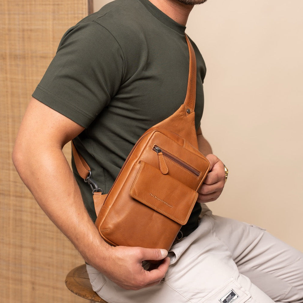 Man holding a cognac leather crossbody bag against a beige wall.