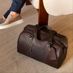 Person sitting on a chair holding a cup with a brown leather duffel bag beside them.