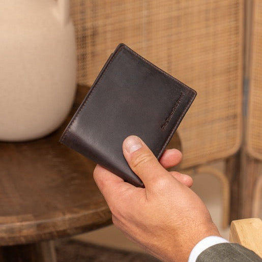 Man holding a brown leather wallet next to a beige vase on a wooden table with a woven wall background.